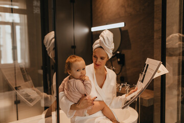 Portrait of funny thoughtful baby in beige blouse and her mother in bathrobe sitting in bathroom and holding newspaper