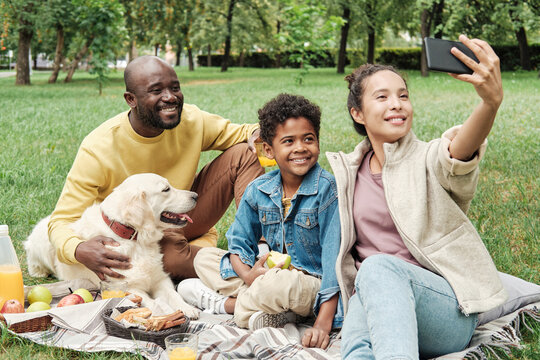 Happy mother holding mobile phone and making selfie portrait of her family with dog on a picnic in the park