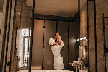 Obraz na płótnie Canvas Lady plays with baby in bathroom. Woman in white bathrobe throws up her little daughter