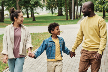 Obraz premium Happy parents holding hands with their child while they walking along the path in the park
