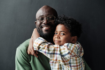 Portrait of African little son embracing his happy father and they smiling at camera isolated on black background