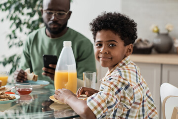 Portrait of African little boy looking at camera while sitting at the table and having breakfast with his family