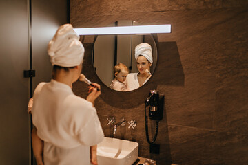 Lady in towel on her head and white bathrobe looks in mirror, holding child in her arms. Woman does makeup in bathroom