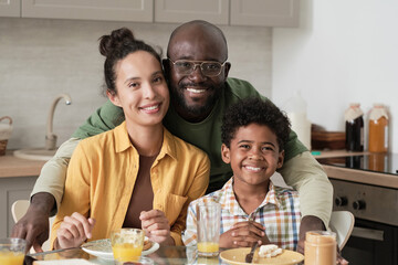 Portrait of African family with little son embracing and smiling at camera while having breakfast at home