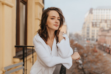 Fototapeta premium Dark-haired young woman in white robe looks at street of city, standing on balcony