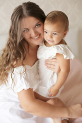 a young beautiful girl with dark long hair in a white dress hugs a small beautiful daughter on a pastel with a pink blanket