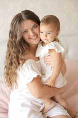 a young beautiful girl with dark long hair in a white dress hugs a small beautiful daughter on a pastel with a pink blanket