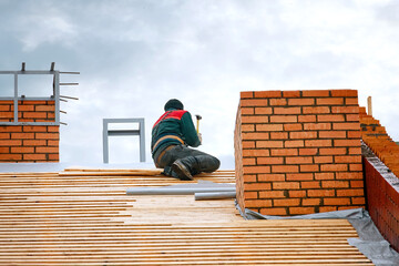 Worker hammers nails with hammer in wooden board on roof at height. Roofer carpenter working on...