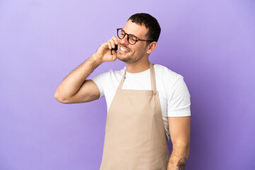 Brazilian restaurant waiter over isolated purple background keeping a conversation with the mobile phone