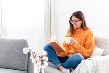 Young woman reading book and drinking coffee at home in autumn. Happy girl wearing in warm sweater relaxing
