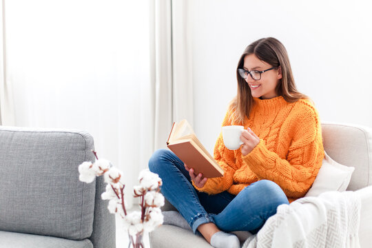 Young woman reading book and drinking coffee at home in autumn. Happy girl wearing in warm sweater relaxing - Powered by Adobe