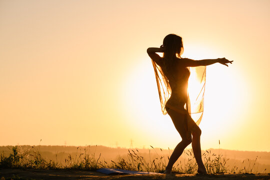 Beautiful girl silhouette dancing exercise against warm sun in transparent cloth, female fit body curves