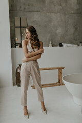 Stylish attractive curly lady in beige trendy jumpsuit and high heels smiles gently, looks down and poses in bathroom.