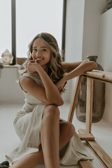 Happy cheerful woman in beige linen dress smiles gently, looks into camera, leans on wooden bench and sits on floor in white cozy bathroom.