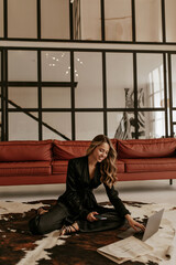 Happy curly woman in stylish black silk suit and sandals sits on carpet on floor in living room, holds phone and works in laptop from home.