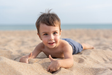 An adorable baby lies on the sand against the background of the sea and smiles at the camera. Recreation