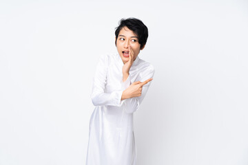 Young Vietnamese woman with short hair wearing a traditional dress over isolated white background pointing to the side to present a product and whispering something