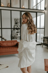 Pretty curly blonde lady in stylish skirt, white blouse puts off jacket and looks into camera in cozy light living room.