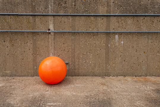 Bright orange bouy hanging from a rope by a concrete wall..