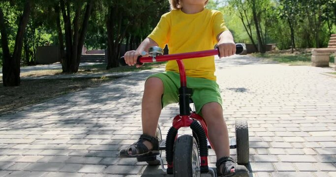Tranquil little boy kid in stylish glasses rides modern red tricycle along road in green city park on sunny summer day closeup