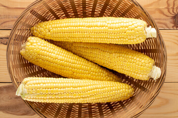 Four cobs of organic sweet corn in a straw dish on a wooden table, close-up, top view.