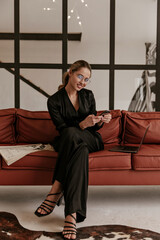 Good-humored woman in trendy black silk jumpsuit and stylish eyeglasses smiles sincerely, holds phone and sits on brown sofa with newspaper and laptop.