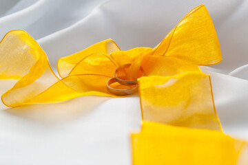 Gold wedding rings and bright yellow ribbon bow on white cloth.