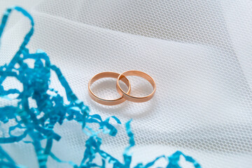 Gold wedding rings on white cloth under a white mesh veil and a blue decorative ribbon. Close-up.