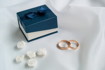 Gold wedding rings, white artificial flowers to decorate the hairstyle and blue jewelry box on white fabric.