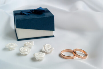 Gold wedding rings, white artificial flowers to decorate the hairstyle and blue jewelry box on white fabric.