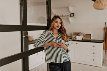 Pretty blonde woman in silk striped shirt holds phone. Curly girl in home outfit leans on window in cozy kitchen.