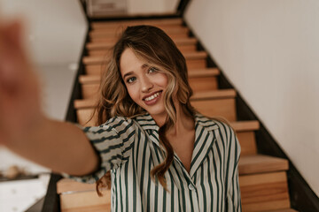 Tanned blue-eyed woman in striped silk green and white shirt smiles sincerely and takes selfie at home on wooden stairs background.