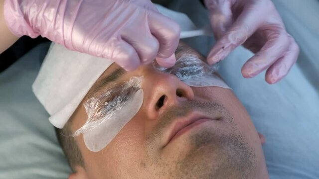 Beautician covered man's lashes with file, pads and towel, lift eyelashes procedure. Man with solution for lamination on lashes, face closeup. Cosmetologist making lash lifting in cosmetology clinic.