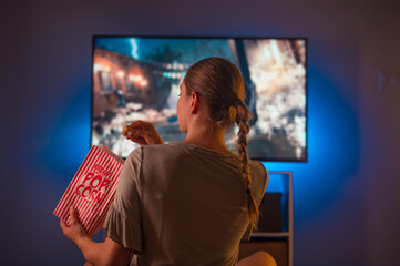 A young blonde girl is watching TV and eating popcorn. Shooting from the back. Color image. Evening time. Blue neon light. Watching movies, TV programs, free time.