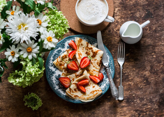 Delicious breakfast, brunch - cappuccino and crepes with strawberries on a wooden table with a bouquet of summer flowers