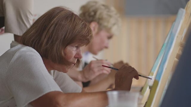 An old retired woman paints a picture together with friends. An elderly group of friends and a senior woman are drawing pictures together.