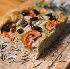 focaccia artesanal con tomates, aceitunas, cebollas en tabla y mesa de madera con harina alrededor