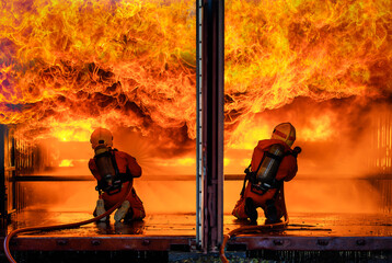 Team of two firefighters wearing full uniform with oxygen tank on back holding pipe spread water and face to wide huge orange hot fire with brave and responsibility in narrow disaster burn area