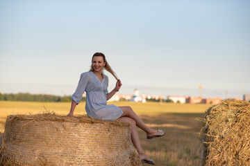 Fototapeta na wymiar Portrait of a young beautiful blonde girl in a light dress on the field near the straw.