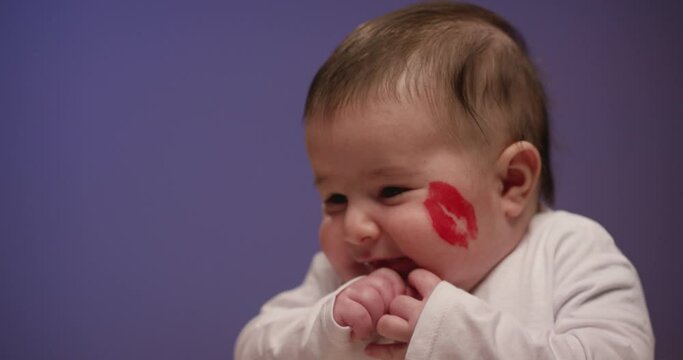 Very funny baby boy with lipstick trail on cheek . Portrait of sweet infant boy with thick cheeks . Little kid smiling and looking around on blue lilac background . Studio shot on RED in slow motion .