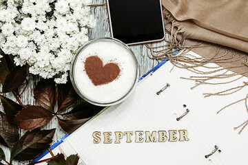 the inscription "September" on a notebook, next to a coffee mug with a cocoa heart, autumn grape leaves and a scarf with a phone. The concept of comfort and relaxation, expectations Flat lay, top view