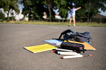 Scattered school supplies and workbooks falling out of an open backpack, lie on the asphalt of the schoolyard against the background of a blurred happy schoolboy raising his hands up