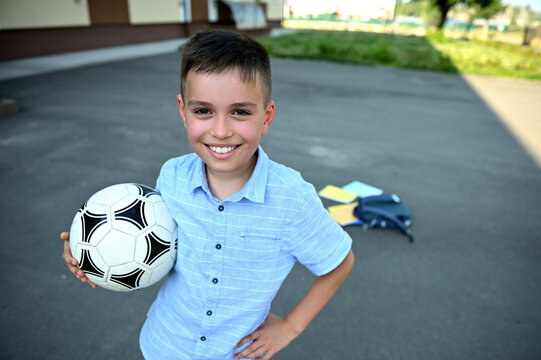 Handsome schoolboy with soccer ball in hands smiling with toothy smile on the background of scattered backpack with school supplies on the schoolyard. Recreation. Rest between lessons
