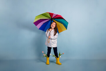 a happy little girl in a raincoat and yellow rubber boots with flowers holds a multi-colored umbrella on a blue background with a copy of the space
