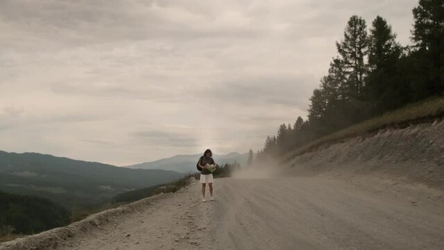 The girl is hitchhiking alone. He walks along the road with a paper map and tries to stop passing cars. Beautiful landscape: dusty road, forest, mountains.
