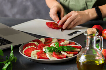 Young woman watching culinary master class online and learning how to cook traditional Italian caprese salad with fresh tomatoes, mozzarella cheese and basil. Home cooking concept.