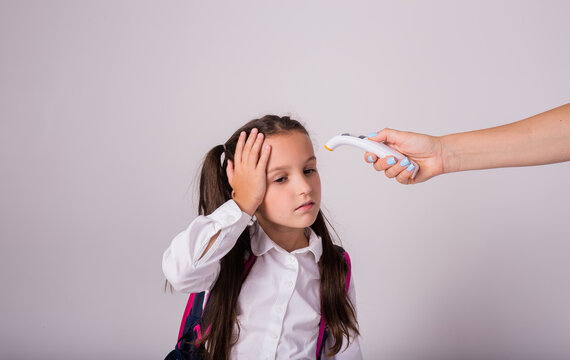 a sick schoolgirl in uniform and with a backpack measures the temperature with an infrared thermometer on a white background with a place for text - Powered by Adobe