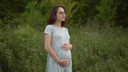 A young pregnant woman stands and holds her belly with her hands. Girl in glasses and a dress in nature.