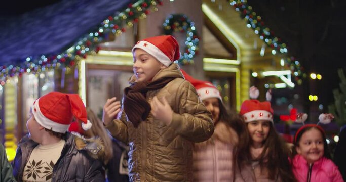 Happy children are jumping , laughing  and looking camera into front of the beautiful house decorated with lights and garlands.  Portrait of many kids with Santa hat and costumes at Christmas night .