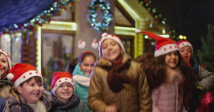 Happy children are jumping , laughing  and looking camera into front of the beautiful house decorated with lights and garlands.  Portrait of many kids with Santa hat and costumes at Christmas night .